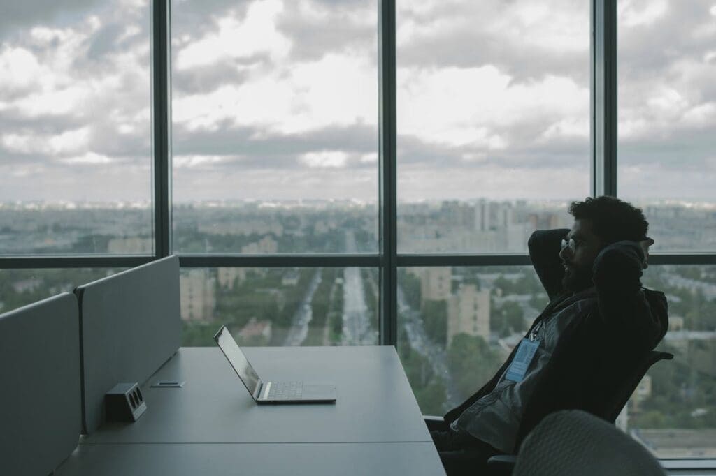 pexels-photo-5483064 A man sits in an office with hands on head in front of a laptop, overlooking a cityscape.
